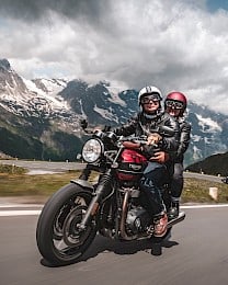 Usměvavý pár na červené motorce Triumph projíždí klikatou horskou silnicí obklopenou zasněženými alpskými štíty a dramatickými mraky. Svoboda a radost z jízdy v srdci hor.