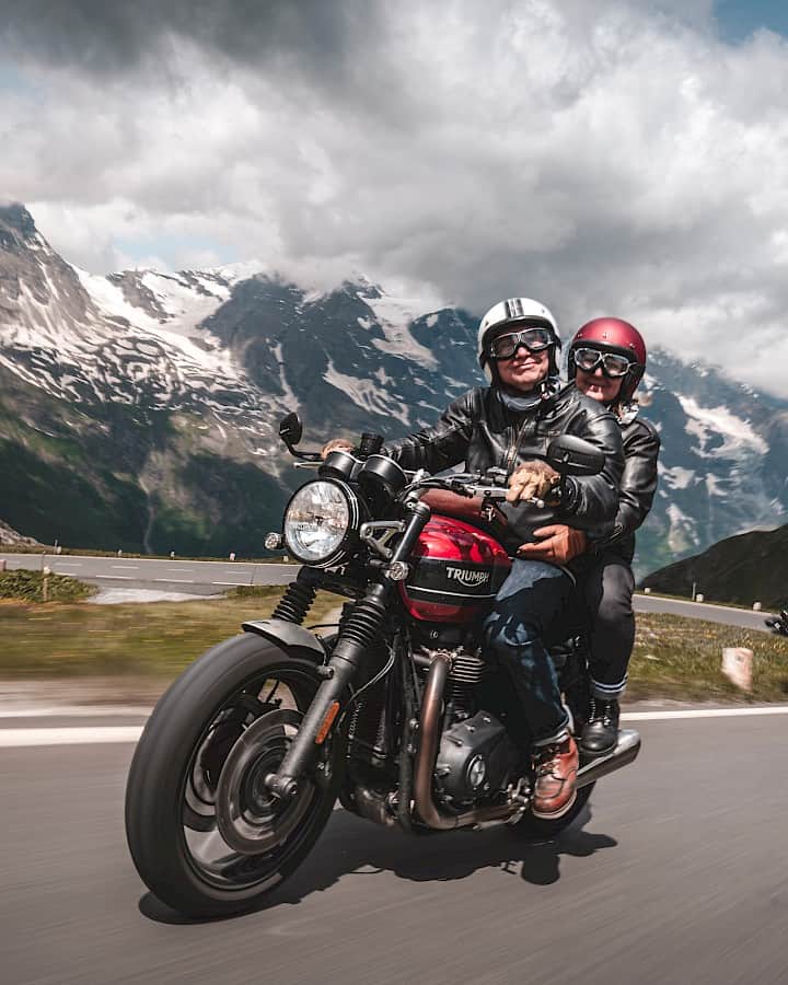 Usměvavý pár na červené motorce Triumph projíždí klikatou horskou silnicí obklopenou zasněženými alpskými štíty a dramatickými mraky. Svoboda a radost z jízdy v srdci hor.