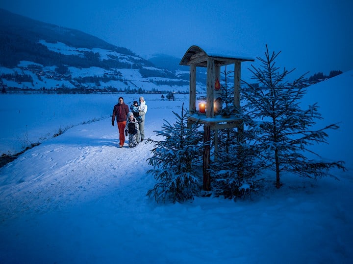 Rodina se prochází po zasněžené stezce se svíčkami ve Wildkogel Areně za soumraku, kolem osvětlené dřevěné lucerny a zimní krajiny.