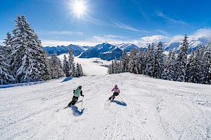 Zwei Skifahrer fahren eine frisch präparierte Piste in der Wildkogel-Arena im Salzburgerland hinunter, umgeben von schneebedeckten Bäumen. Die Sonne scheint strahlend vom blauen Himmel, während sich im Tal unterhalb Nebel sammelt. Im Hintergrund erheben sich beeindruckende, verschneite Alpenberge.