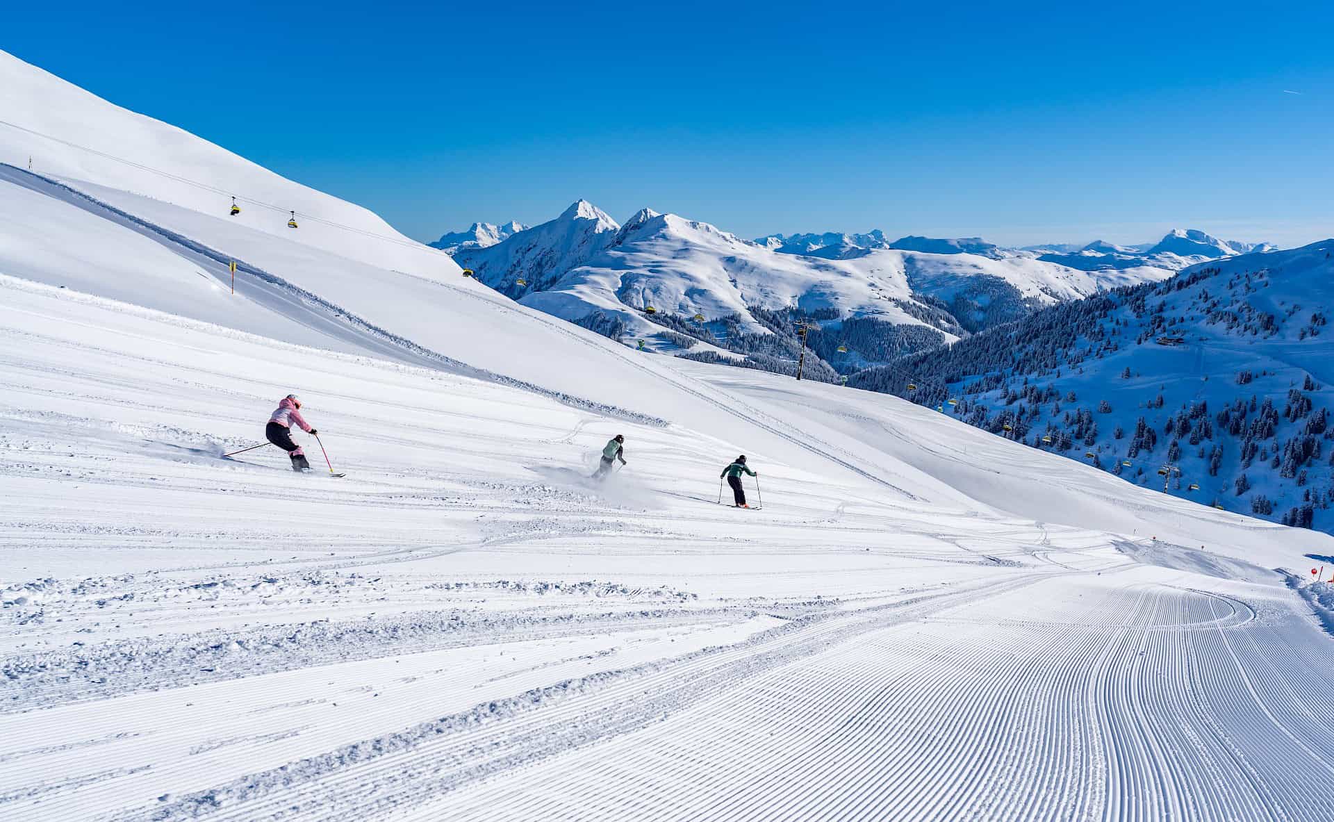 Lyžaři si užívají perfektně upravené sjezdovky v oblasti Wildkogel Arena – slunečné zimní počasí a nádherný výhled na zasněžené hory v ceně.
