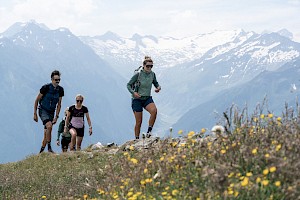 Eine sportliche Wandergruppe erklimmt einen Bergpfad im Höhenwandergebiet Wildkogel mit Blick auf die verschneiten Gipfel der österreichischen Alpen – aktiver Sommerurlaub am Hohe Tauern Panorama Trail inmitten atemberaubender Natur.