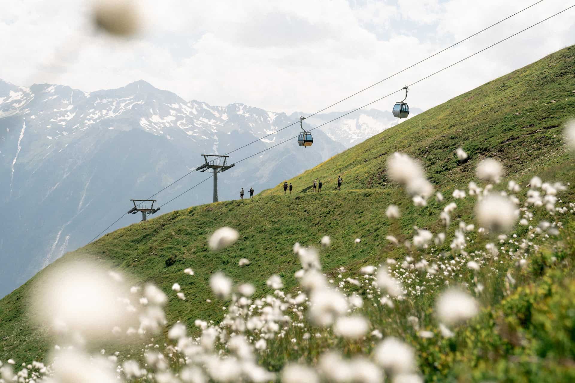 Turisté na rozkvetlé alpské stezce ve vysokohorském turistickém regionu Wildkogel s výhledem na lanovku a zasněžené vrcholy Vysokých Taur – letní dovolená na stezce Hohe Tauern Panorama Trail uprostřed květinové alpské krajiny a impozantních hor.