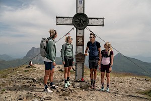 Vier Wanderer erreichen lachend den Gipfel in der Wildkogel-Wildkogel-Arena und versammeln sich am imposanten Gipfelkreuz mit Blick auf den Großen Rettenstein – unvergesslicher Wandermoment im Salzburger Land.