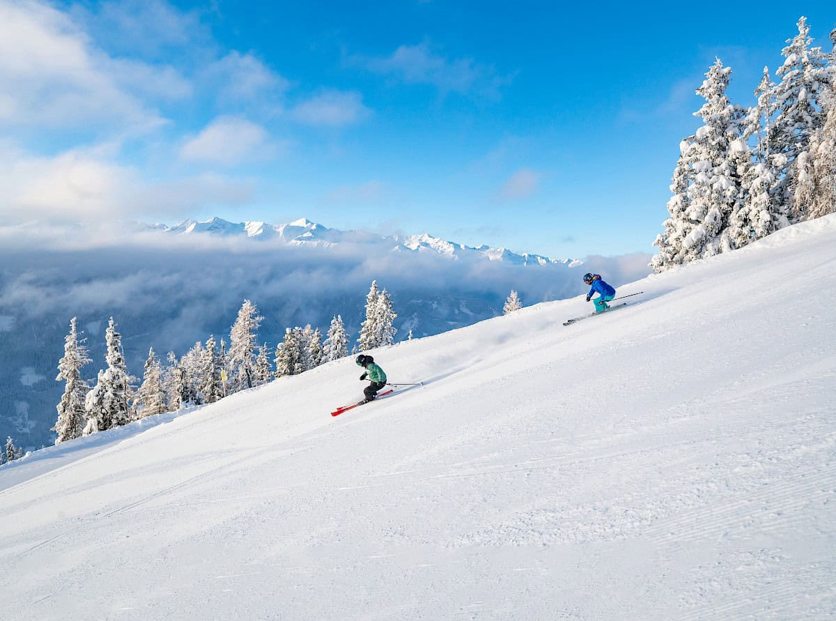 Dva lyžaři svižně projíždějí čerstvým prašanem v oblasti Wildkogel – obklopeni zasněženými stromy a majestátními vrcholy Vysokých Taur v pozadí.