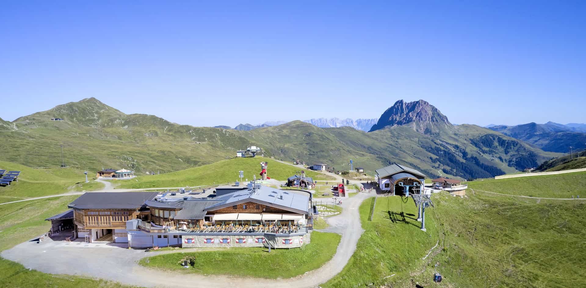 Panoramatický pohled na horskou restauraci u horní stanice lanovky Wildkogel v létě, obklopenou zelenými pastvinami a turistickými stezkami – v pozadí výrazný vrchol Großer Rettenstein.