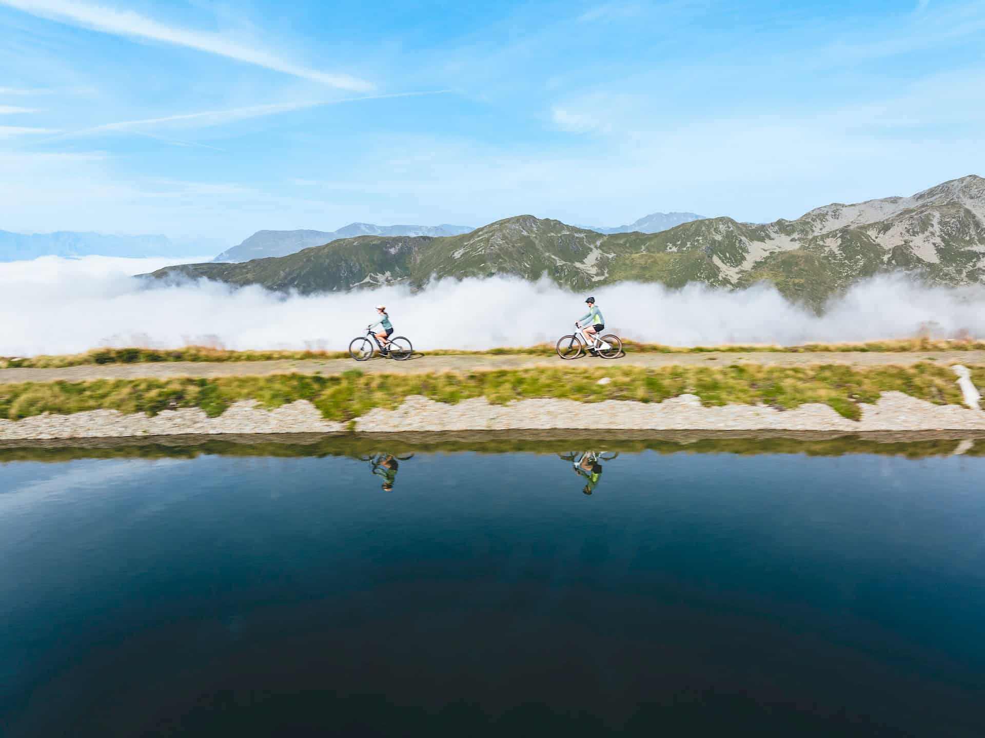 Dva cyklisté si užívají panoramatickou jízdu podél čistého horského jezera s výhledem na Vysoké Taury ve Wildkogel Areně Neukirchen & Bramberg.