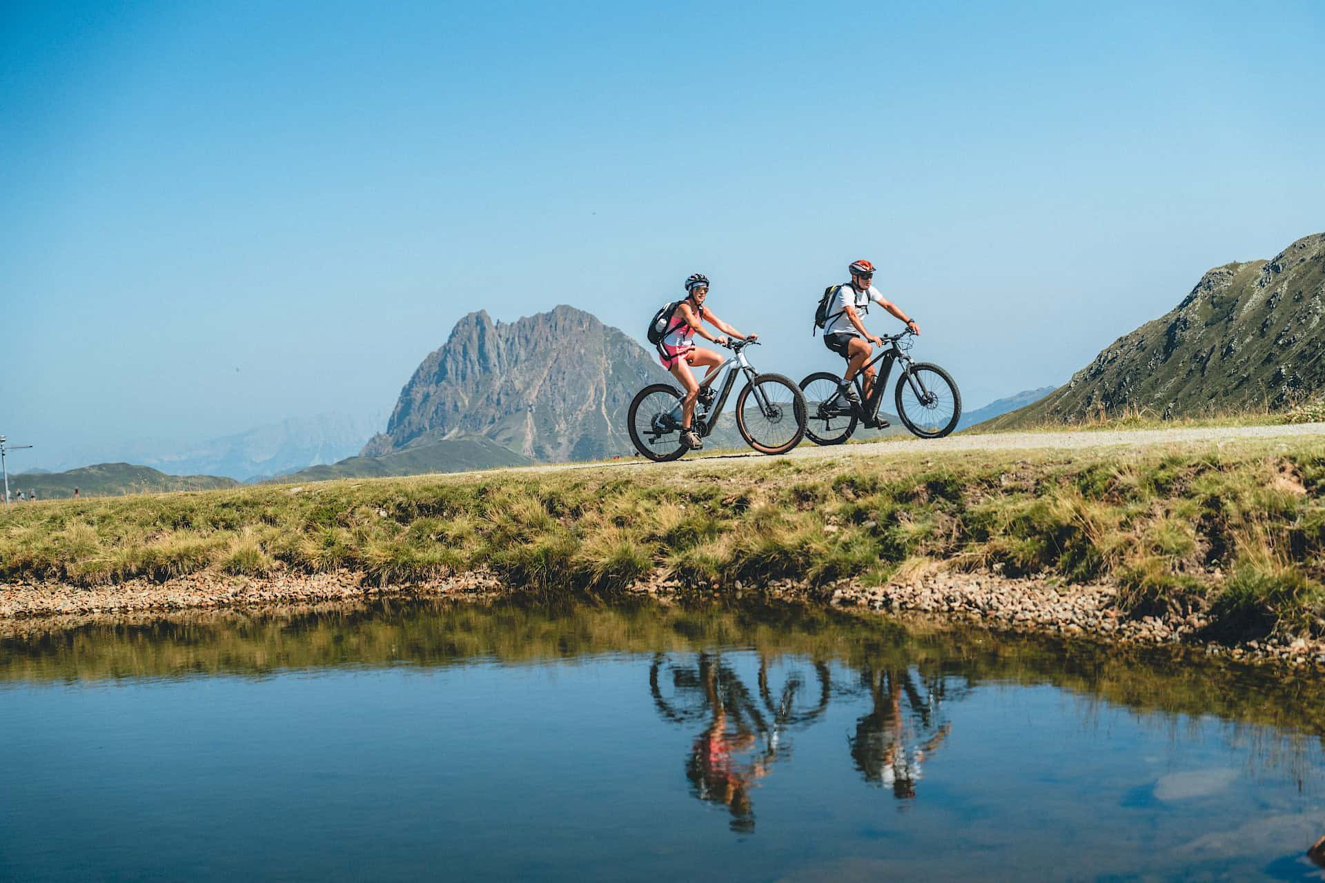 Dva cyklisté na elektrokolech si užívají panoramatickou jízdu v horské oblasti Wildkogel – aktivní letní zážitky v pohoří Vysoké Taury s výhledem na majestátní Großer Rettenstein a křišťálově čistou vodou v popředí.