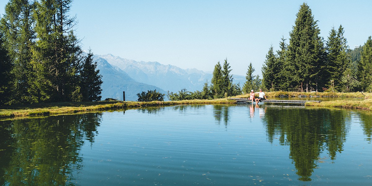 Zwei Wanderer genießen eine entspannte Pause am idyllischen Speicherteich im Höhenwandergebiet Wildkogel, umgeben von grüner Bergnatur und einem beeindruckenden Panoramablick auf die Hohen Tauern – pures Naturerlebnis im Bergsommer. uk-cover