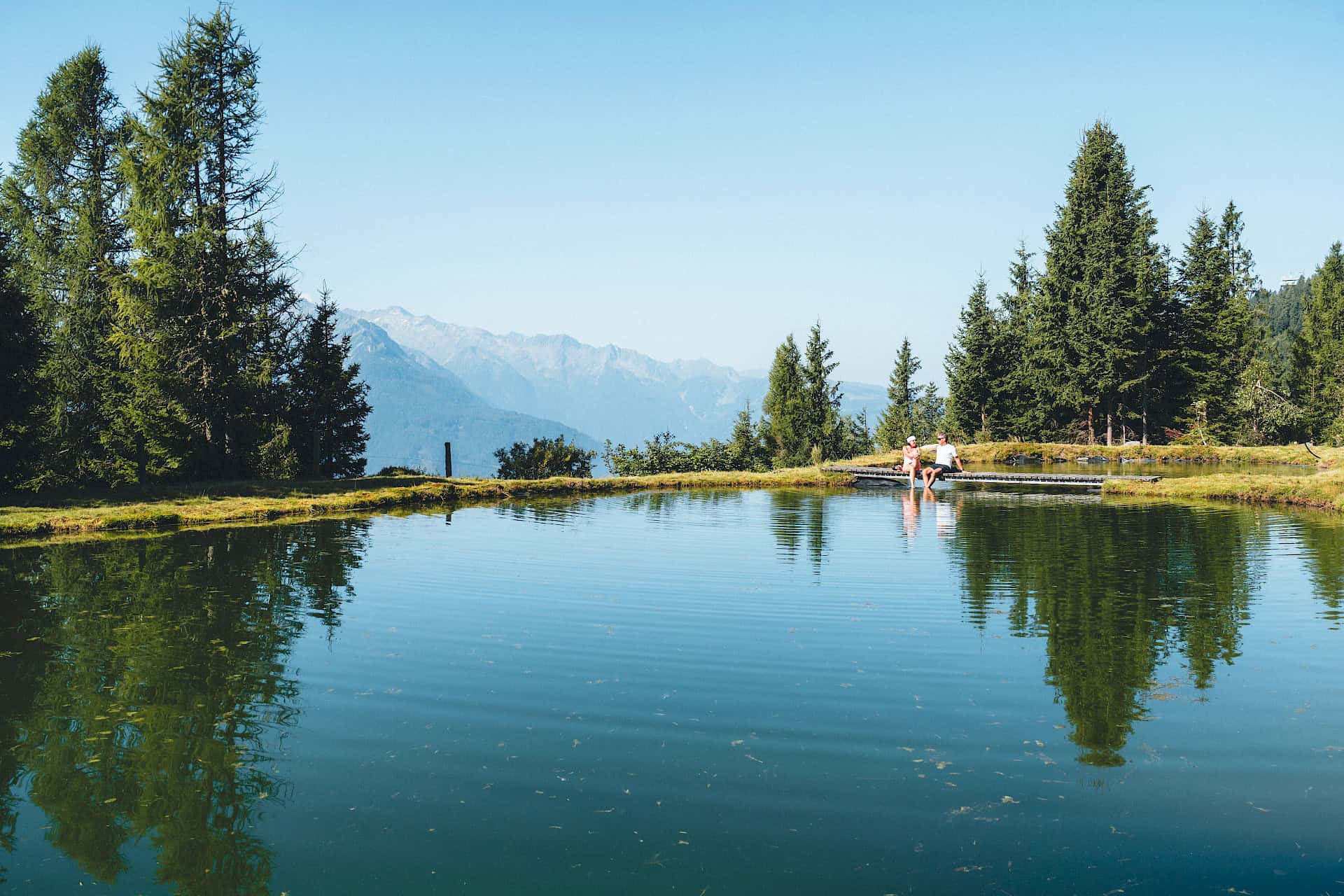 Dva turisté si užívají klidnou pauzu u idylického jezírka v horské oblasti Wildkogel, obklopeni zelenou alpskou přírodou a nádherným výhledem na Vysoké Taury – čistý přírodní zážitek během letní turistiky v horách.