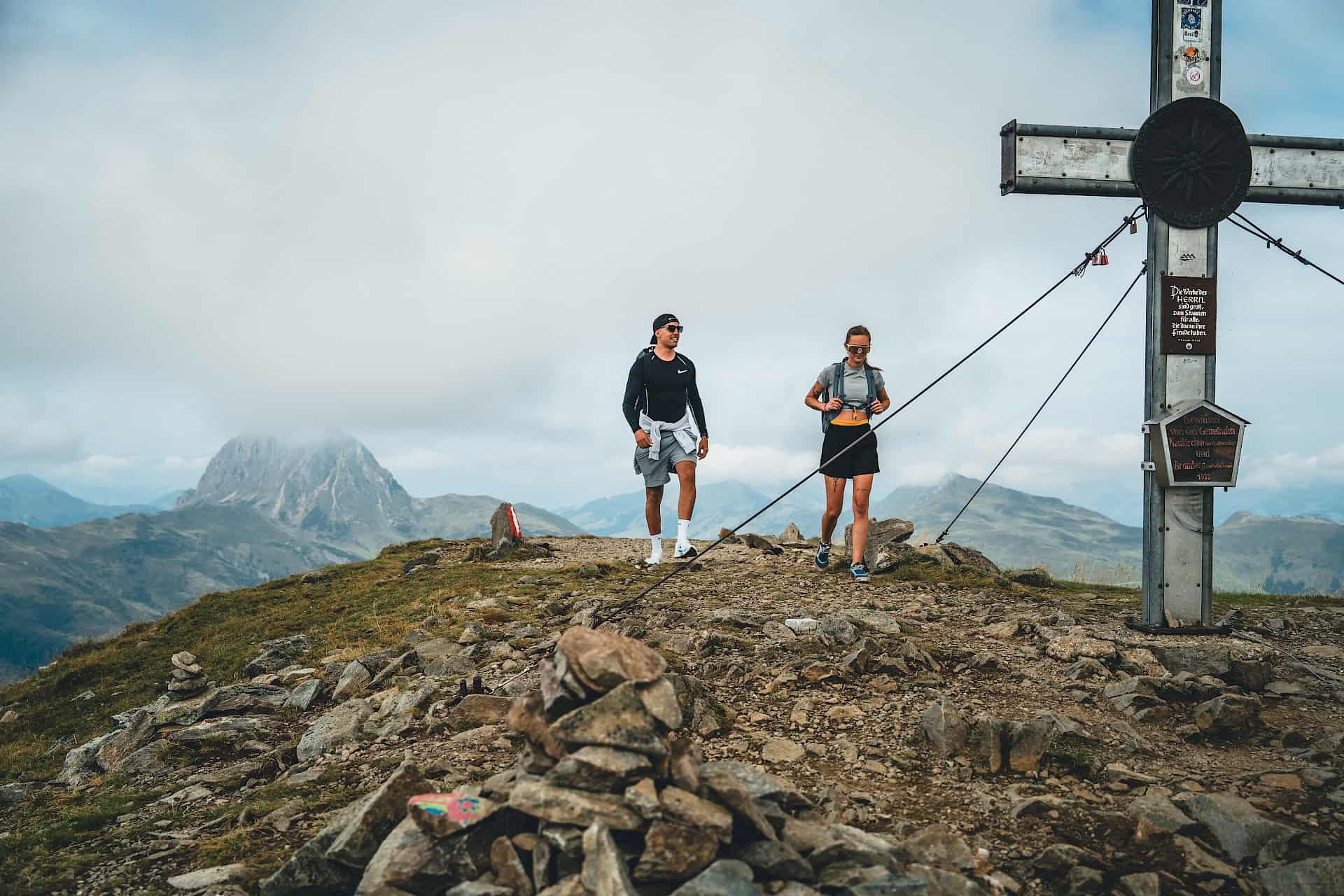 Dva turisté dorazí ke kříži na vrcholu ve Wildkogel Areně, v pozadí je vidět oblačný Großer Rettenstein.