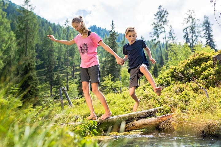 Zwei Kinder balancieren barfuß über einen Baumstamm am Wasserlauf in der Wildkogel-Arena – naturnahes Abenteuer und spielerisches Entdecken beim Familienurlaub in den österreichischen Alpen.
