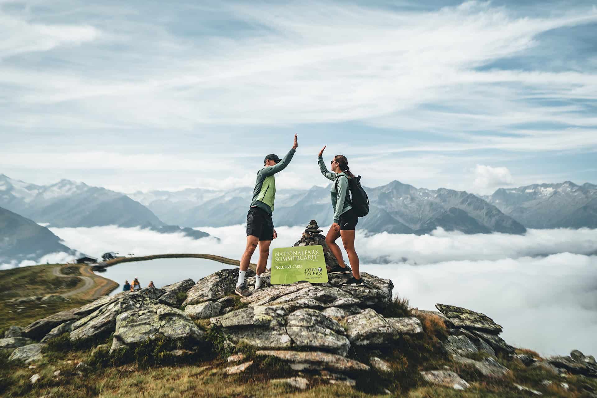 Zwei Wanderer geben sich ein High-Five am Gipfel in der Wildkogel-Arena neben einem Schild der Nationalpark Sommercard – Bergmoment mit Blick auf die Hohen Tauern und inklusive Urlaubsvorteilen.