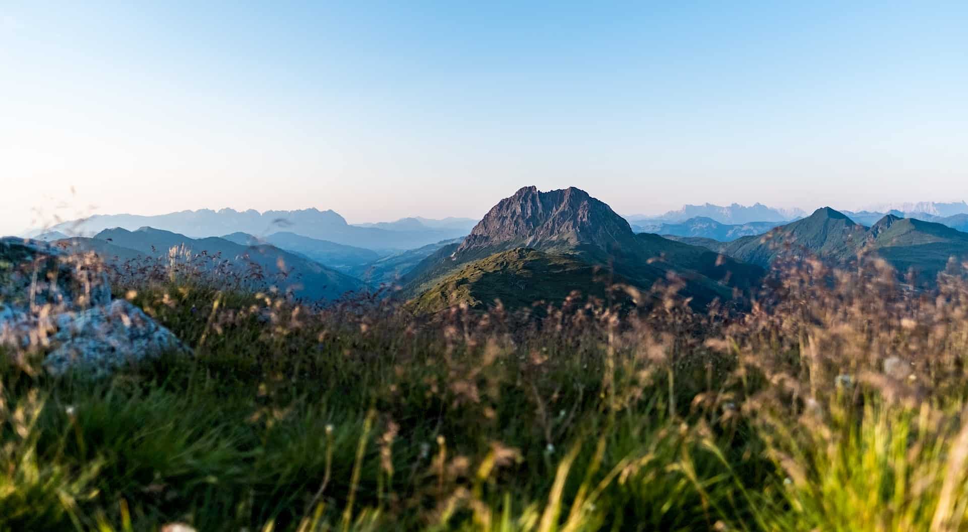 The Großer Rettenstein stands out prominently in the summer alpine landscape, surrounded by gentle green hills and blooming alpine meadows under a clear blue sky.