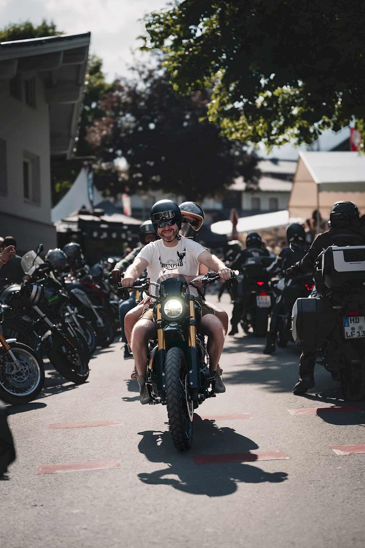 Motorrijders op het motorfestival The Newchurch Summit in een dorpssetting: Een lachende bestuurder met een passagier op een moderne café-racer rijdt door een steeg, omringd door andere bikers en geparkeerde motorfietsen – zomerse sfeer met festivalsfeer.