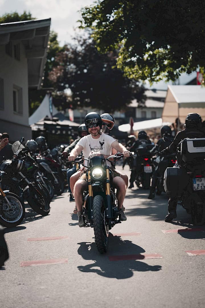 Motorrijders op het motorfestival The Newchurch Summit in een dorpssetting: Een lachende bestuurder met een passagier op een moderne café-racer rijdt door een steeg, omringd door andere bikers en geparkeerde motorfietsen – zomerse sfeer met festivalsfeer.