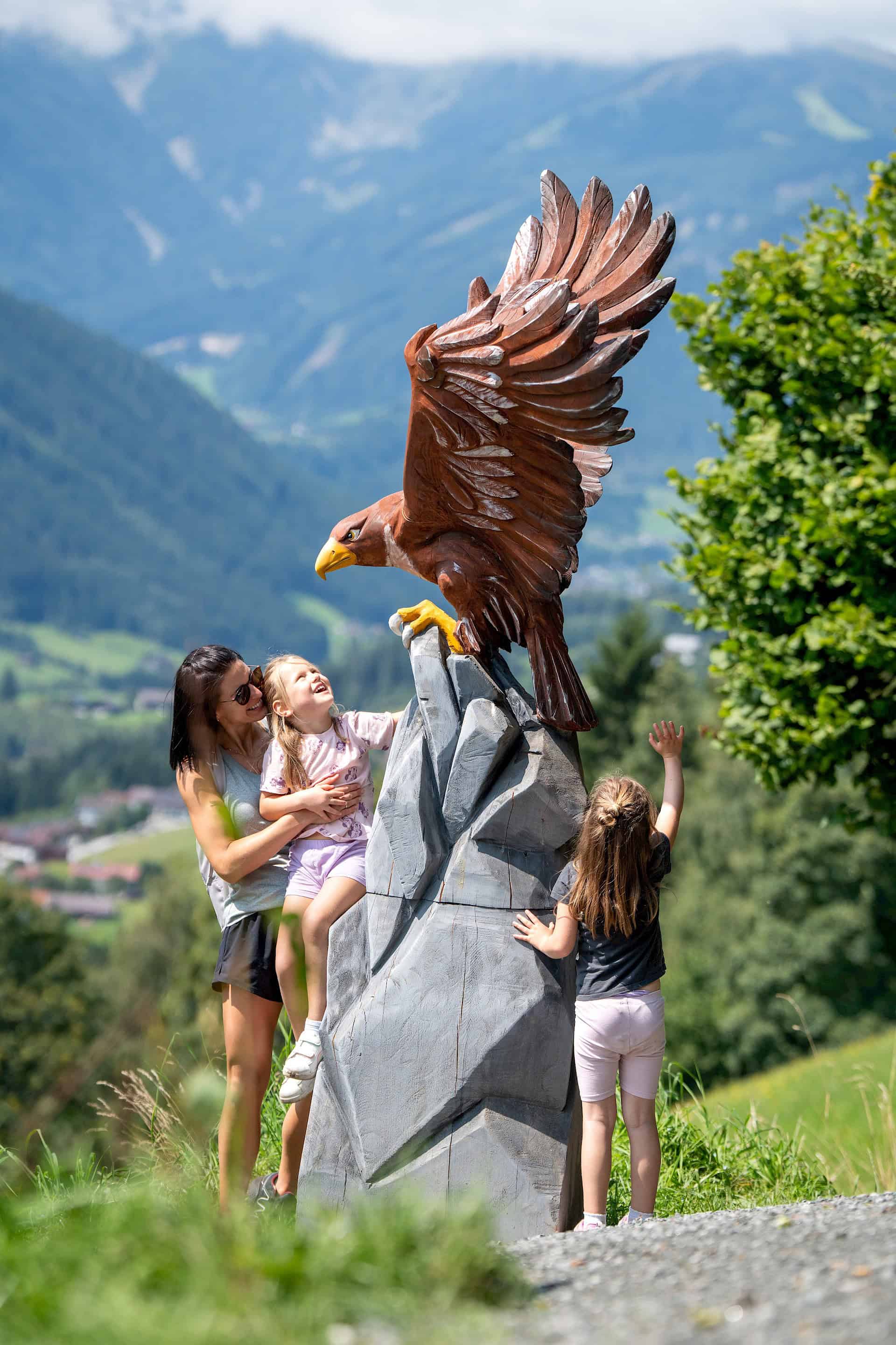 Maminka s dcerami obdivují velkou dřevěnou sochu orla na zážitkové stezce „Adler Toni Weg“ v oblasti Wildkogel Arena.