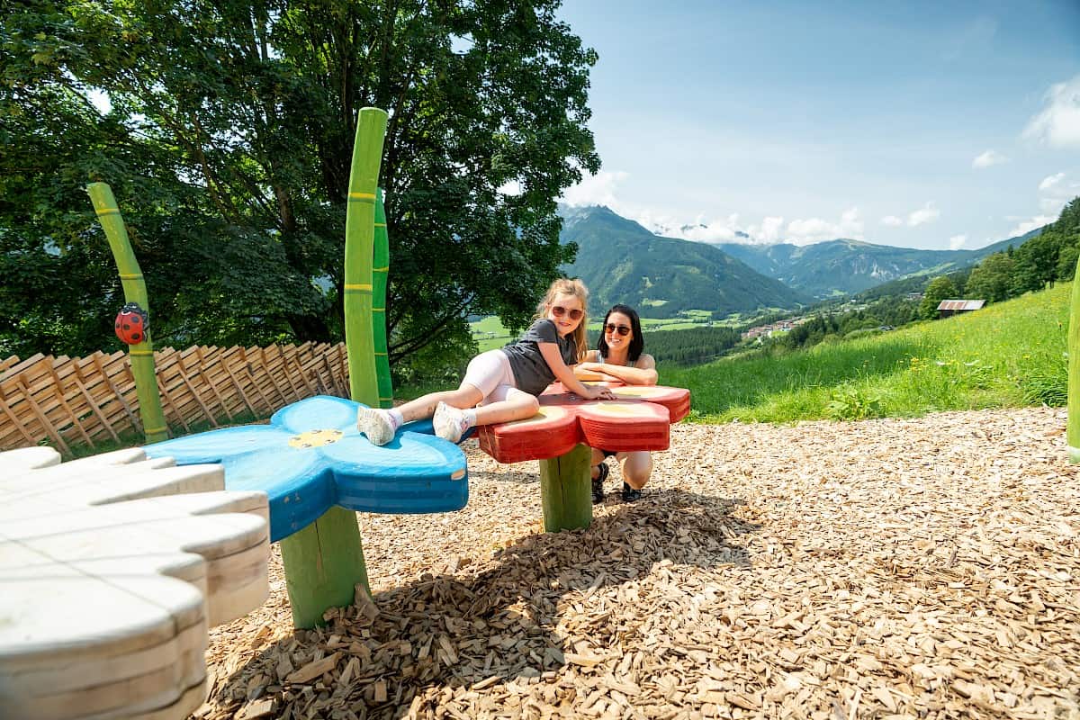 Mother and daughter explore a colorful flower-shaped play station along the "Adler Toni Trail", overlooking the lush valley and surrounding mountains.