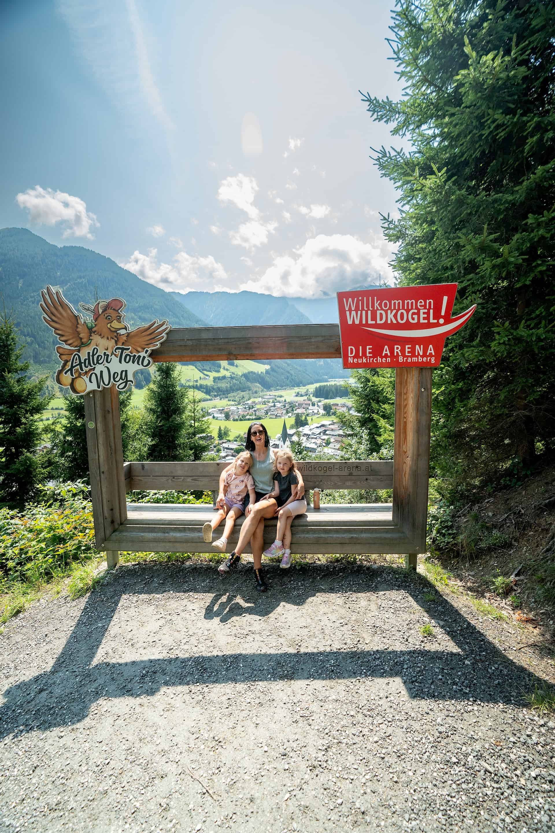Matka s dcerami si dávají pauzu na velké dřevěné lavičce s výhledem do údolí na zážitkové stezce „Adler Toni Weg“ v oblasti Wildkogel-Arena.