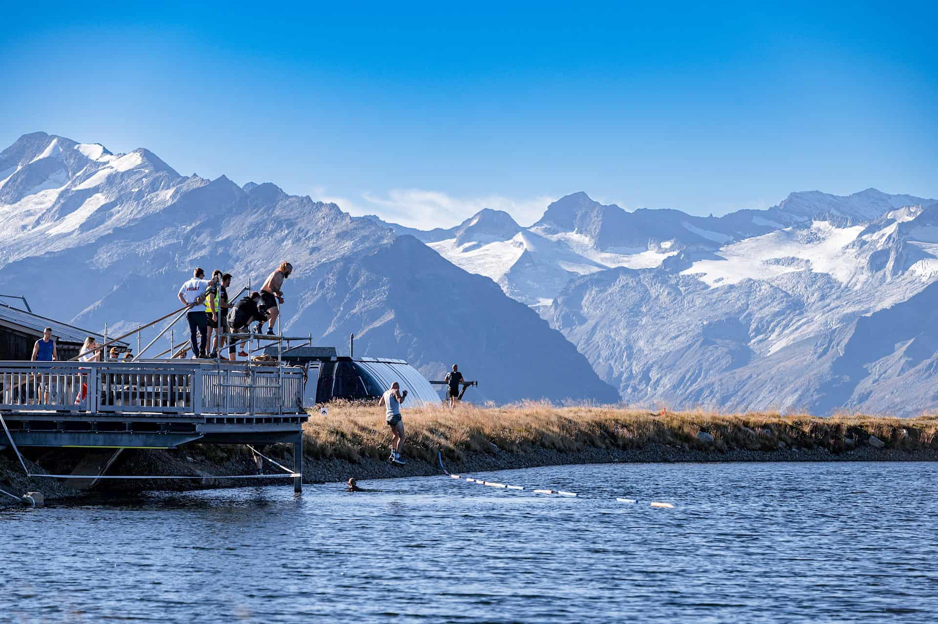 Teilnehmende des Predator Race springen von einer Plattform in den Bergsee vor beeindruckender Hochgebirgskulisse.