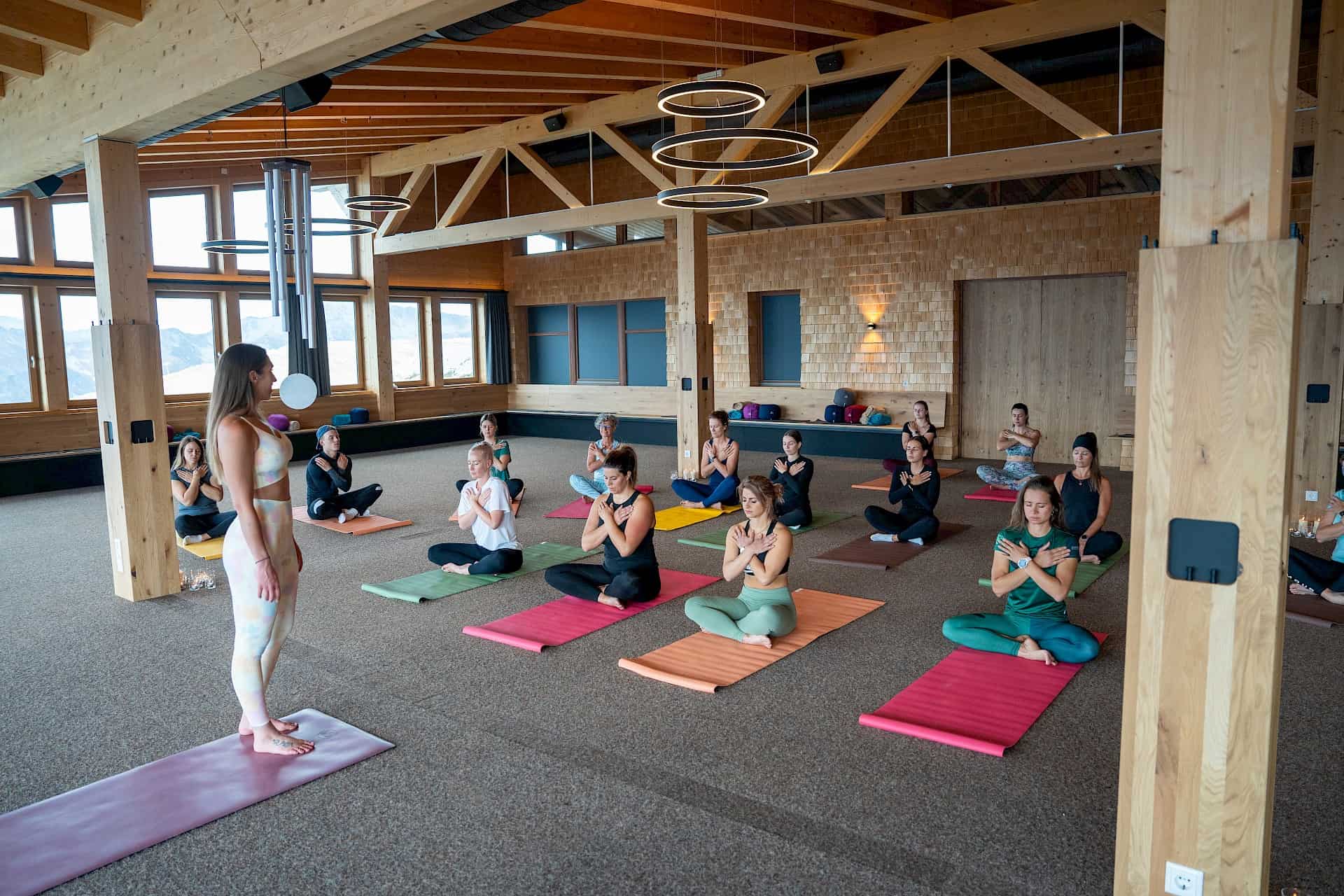 Innenraumaufnahme einer Yogaeinheit während des "Bergzeit – move & soul retreat". Die Teilnehmer:innen sitzen in stiller Achtsamkeit mit überkreuzten Armen auf bunten Yogamatten in einem lichtdurchfluteten Raum mit alpinem Holzdesign.
