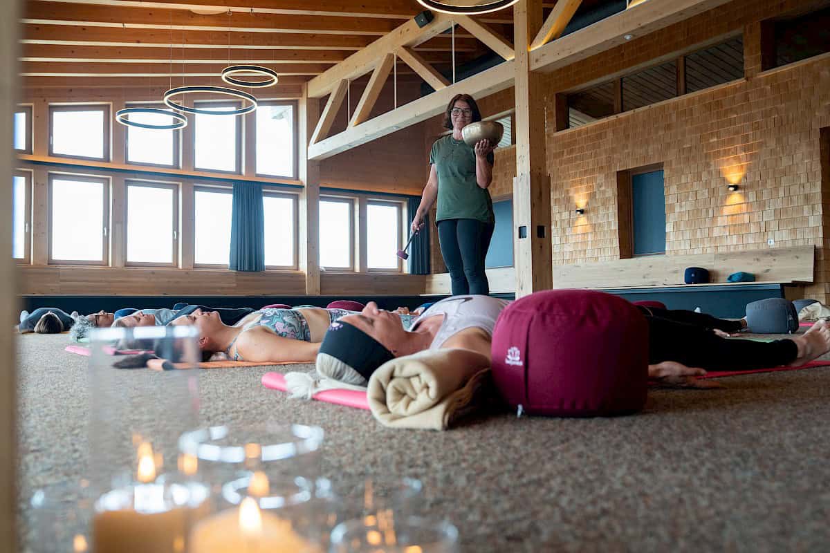 Innenaufnahme einer geführten Entspannungsübung beim „Bergzeit – move & soul retreat“. Teilnehmer:innen liegen in Savasana auf Yogamatten, während Trainerin Ulli mit einer Klangschale durch den Raum geht. Im Vordergrund brennen stimmungsvolle Kerzen.