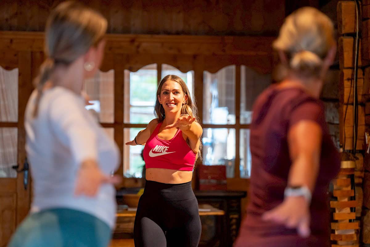 Trainerin Nina leitet eine Yogaeinheit im Innenbereich eines gemütlichen Holzhauses. Im Vordergrund sieht man Teilnehmerinnen in der Yoga-Haltung „Krieger II“, während Nina mit strahlendem Lächeln motivierend anleitet.