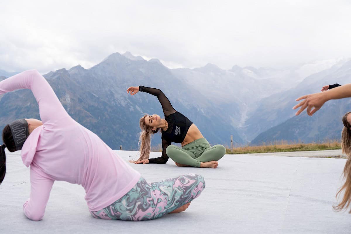 Trainerin Ewi führt eine Yogaeinheit auf einer Panorama-Plattform am Wildkogel mit Blick ins Habachtal an. In sitzender Seitdehnung demonstriert sie Eleganz und Achtsamkeit vor einer eindrucksvollen Alpenkulisse.