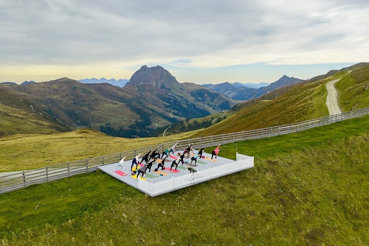 Yogales op een vrijstaande platform hoog boven het dal, omgeven door alpenweiden. Op de achtergrond steekt de markante top van de Großer Rettenstein trots omhoog.