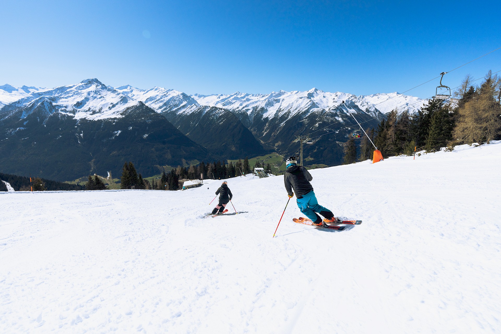 Twee skiërs dalen dynamisch af op een brede, zonnige piste met een adembenemend uitzicht op de met sneeuw bedekte drieduizenders van de Hohe Tauern.