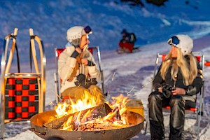 Zwei Personen sitzen nach dem Rodeln gemütlich an einer Feuerschale und wärmen sich im Schein des Feuers auf der verschneiten Rodelbahn.