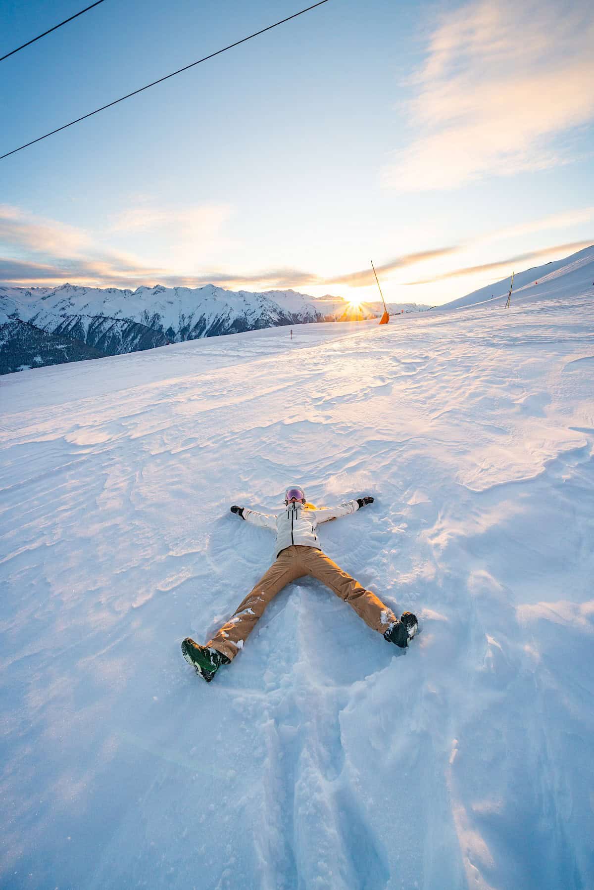 Person liegt im Schnee und formt einen Schneeengel, mit Blick auf den Sonnenuntergang über dem Wildkogel-Skigebiet