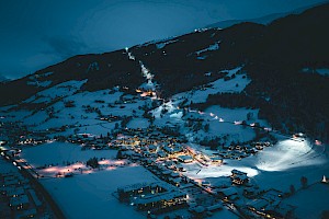 Nächtliche Winteraufnahme der Wildkogel-Arena mit beleuchteten Häusern im Tal und der hell erleuchteten Skipiste am Berg.