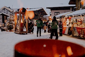 Winterlicher Adventmarkt mit verschneiten Ständen, Besuchern in warmen Jacken und einem unscharfen Feuerkorb im Vordergrund, der für stimmungsvolle Atmosphäre sorgt.