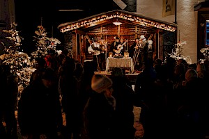 Stimmungsvolle Adventszene mit Musikanten auf einer warm beleuchteten Holzhütte und zahlreichen Besuchern, die im winterlichen Abendlicht zuhören.