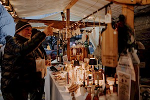Besucher betrachtet handgemachte Produkte an einem festlich beleuchteten Adventmarktstand, umgeben von warmem Lichterglanz und winterlicher Stimmung.