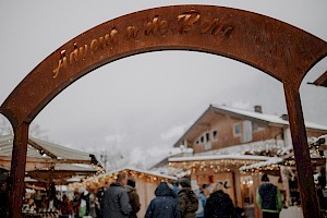 Eingangsportal „Advent a de Berg“ aus Metall, dahinter der winterlich verschneite Adventmarkt mit Besucherinnen und Besuchern sowie festlich beleuchteten Ständen.