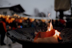 Brennende Feuerschale im Vordergrund, dahinter unscharfe Lichter des Adventmarkts, die eine warme und gemütliche Winterstimmung erzeugen.
