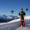 Mann mit Rodel in der Hand und im Hintergrund ist die Smaragdbahn der Wildkogel-Arena zu sehen.