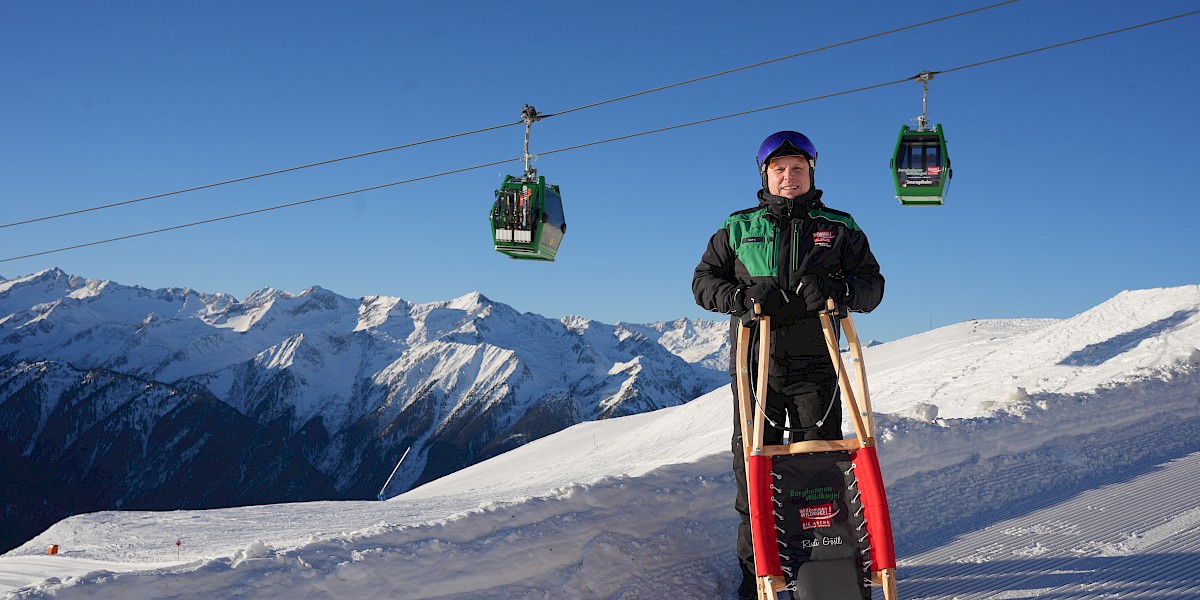 Mann mit Rodel in der Hand und im Hintergrund ist die Smaragdbahn der Wildkogel-Arena zu sehen. uk-cover