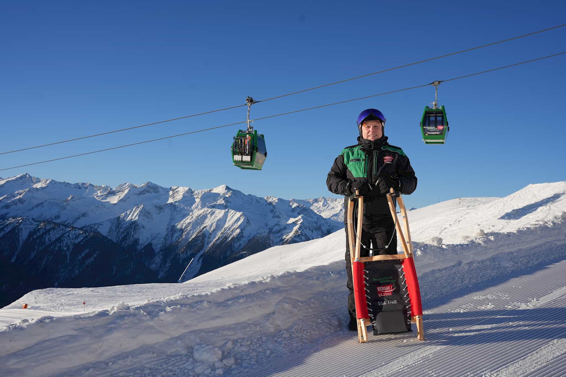 Mann mit Rodel in der Hand und im Hintergrund ist die Smaragdbahn der Wildkogel-Arena zu sehen.