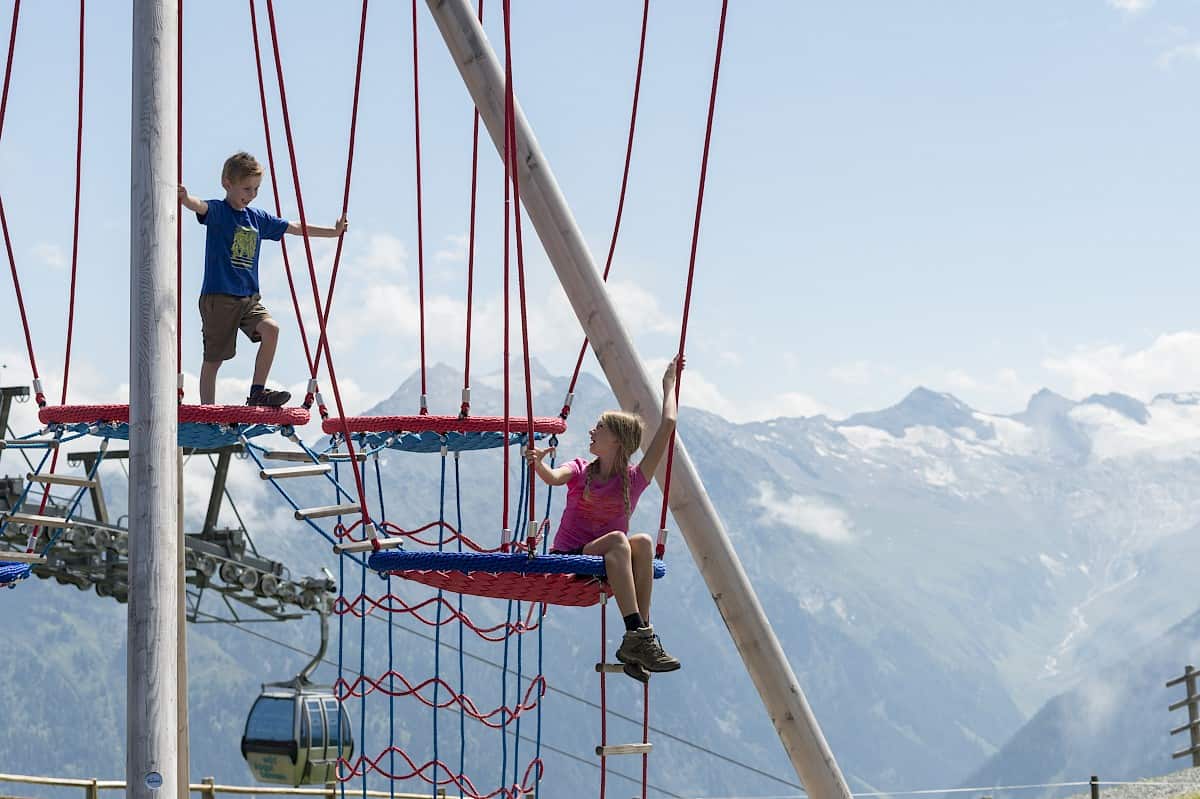 Children climbing in the Kogel-Mogel Adventure Arena in the Wildkogel Arena, Austria – outdoor activities for families with children during a summer holiday in the Alps with views of the 3,000-metre peaks of the Hohe Tauern National Park.