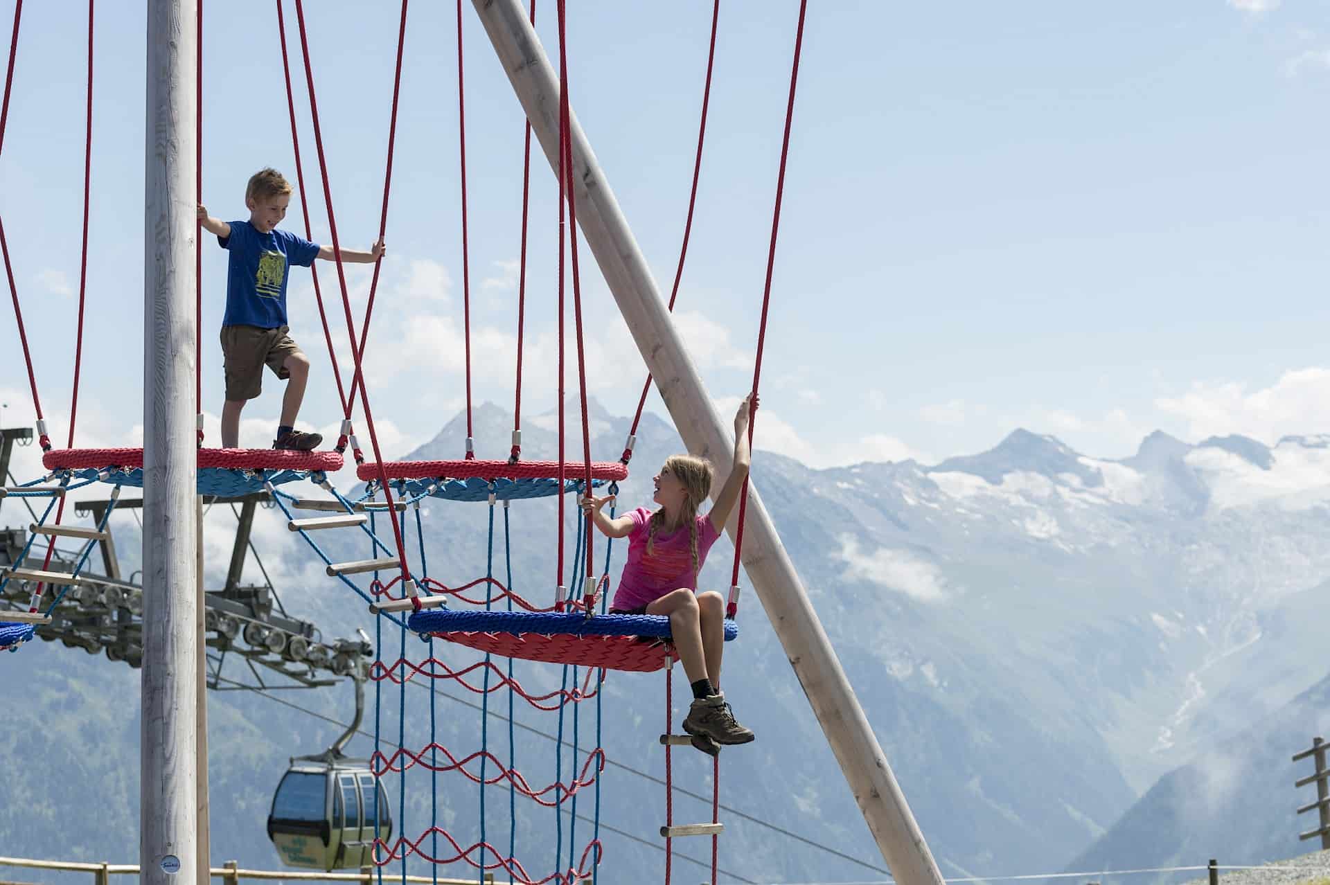 Children climbing in the Kogel-Mogel Adventure Arena in the Wildkogel Arena, Austria – outdoor activities for families with children during a summer holiday in the Alps with views of the 3,000-metre peaks of the Hohe Tauern National Park.