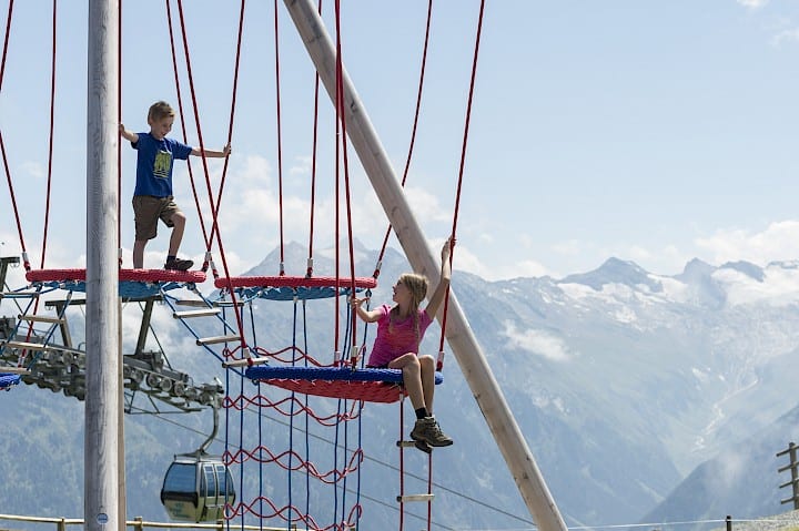 Kinder beim Klettern in der Abenteuer-Arena Kogel-Mogel in der Wildkogel-Arena in Österreich – Outdoor-Aktivitäten für Familien mit Kindern im Sommerurlaub in den Alpen mit Blick auf die 3.000er des Nationalparks Hohe Tauern.