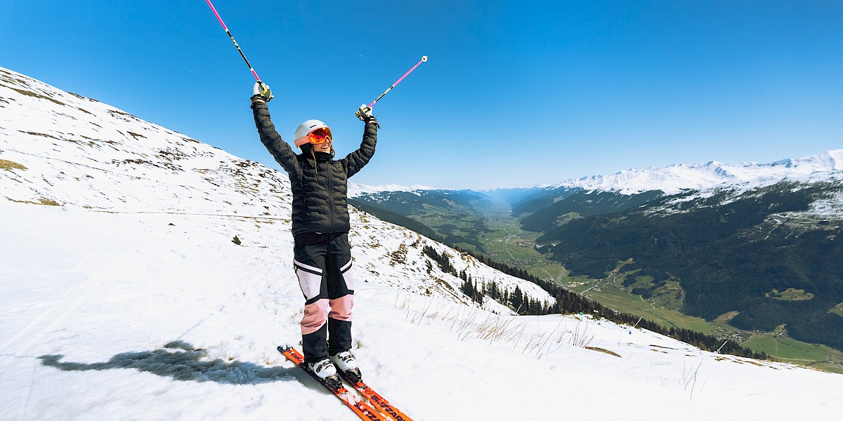 Skifahrer hebt jubelnd die Stöcke beim Sprung im Skigebiet, strahlender Sonnenschein und weiter Blick über die verschneiten Alpen. uk-cover