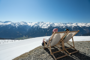Zwei Personen entspannen in Liegestühlen auf einem Aussichtspunkt im Skigebiet, vor ihnen ein Panorama mit verschneiten Alpen