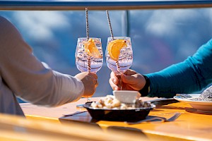 Zwei Personen stoßen mit Aperitifgläsern auf einer Sonnenterrasse im Skigebiet an, im Vordergrund ein Tisch mit Essen.
