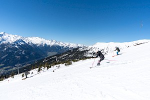 Zwei Skifahrer fahren auf einer breiten Piste bei Sonnenschein, im Hintergrund ein Panorama mit verschneiten Alpen.