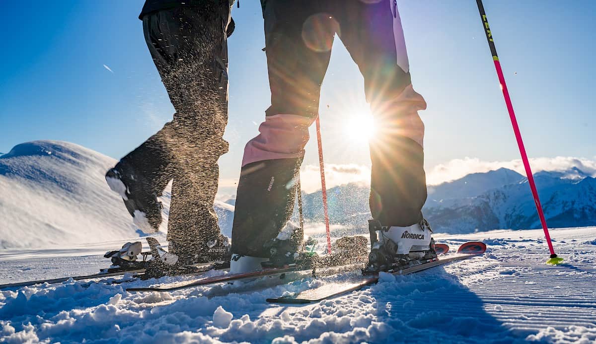 Nahaufnahme von Skischuhen im Schnee mit Sonnenstrahlen dazwischen, im Hintergrund verschneite Berge.