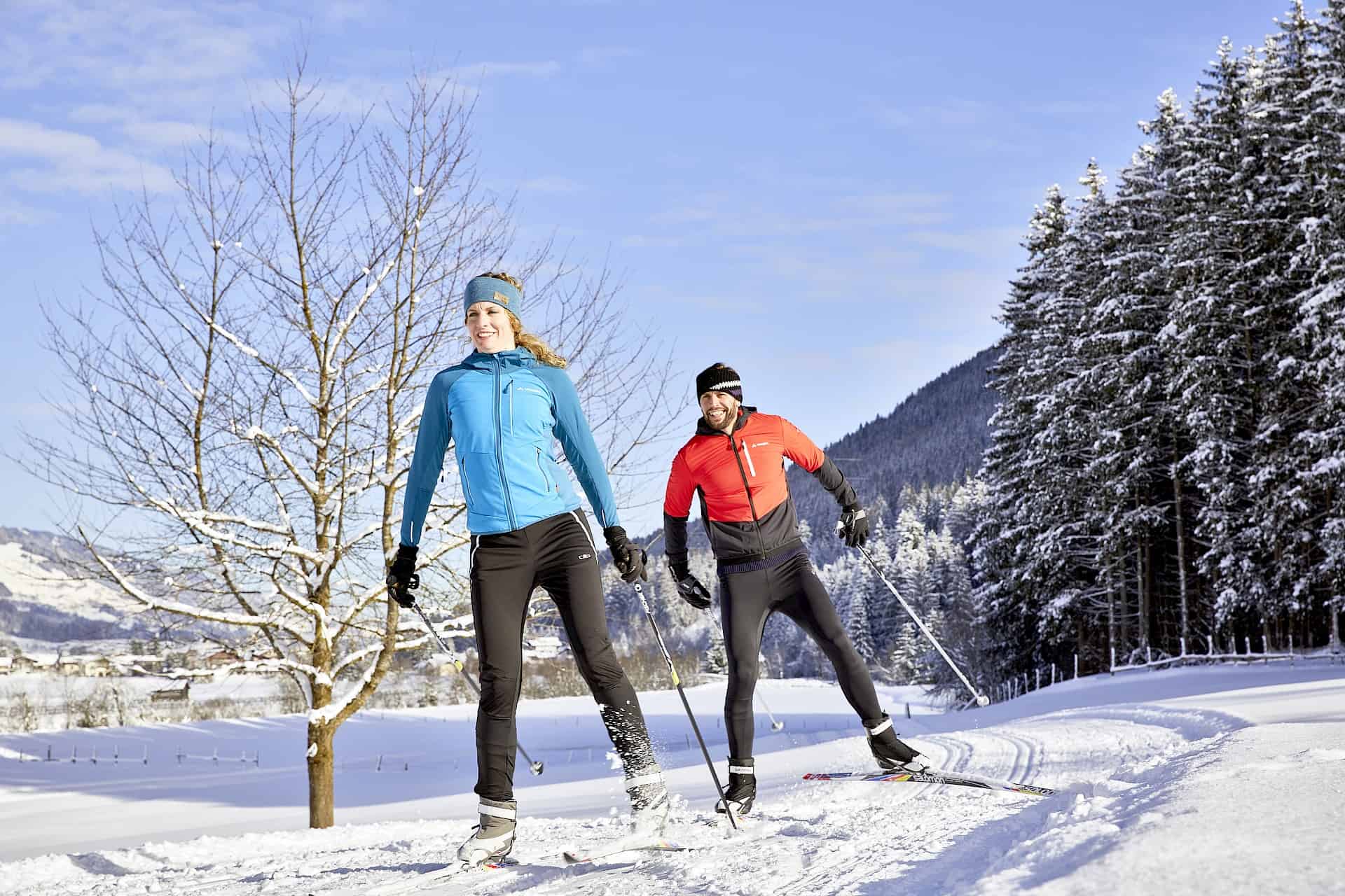 Ein sportliches Paar beim Langlaufen auf frisch gespurter Loipe in der Ferienregion Nationalpark Hohe Tauern bei Bramberg am Wildkogel – umgeben von verschneiten Wäldern und traumhafter Winterlandschaft im Sonnenschein. Aktiver Winterurlaub in der Wildkogel-Arena.